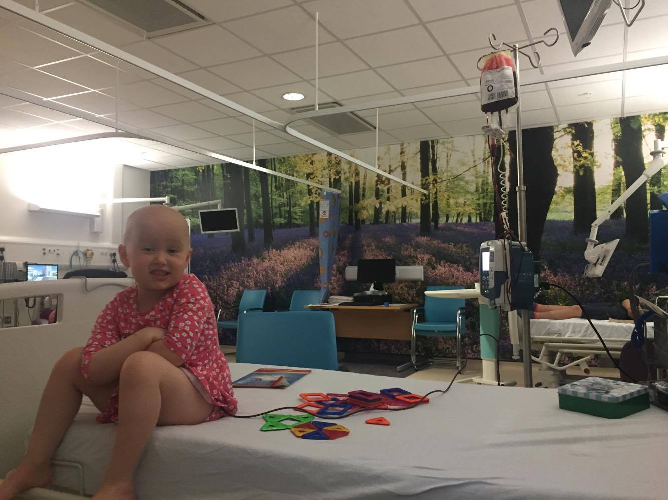 Emily, a small girl with no hair after chemotherapy, smiles at the camera while having a blood transfusion in hospital.