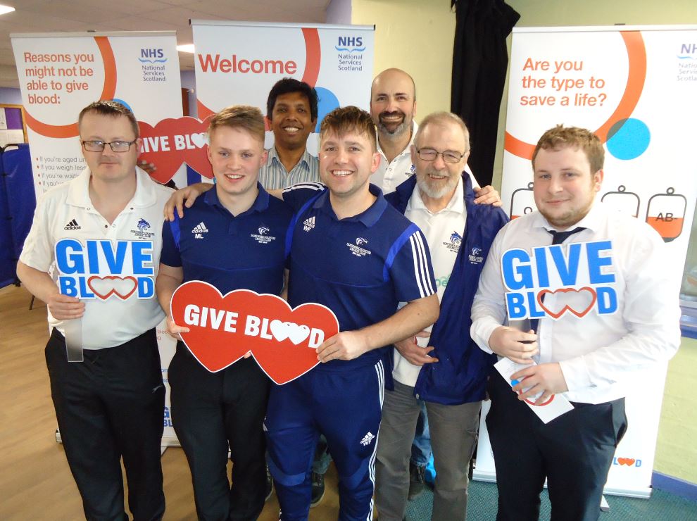 Members of Northern Counties Cricket Club smile and pose for photo while holding Give Blood signs. 