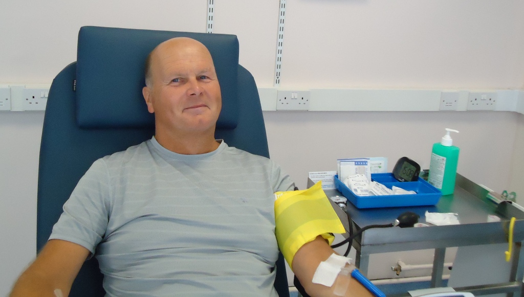 Paul sits on bed smiling after giving blood. 