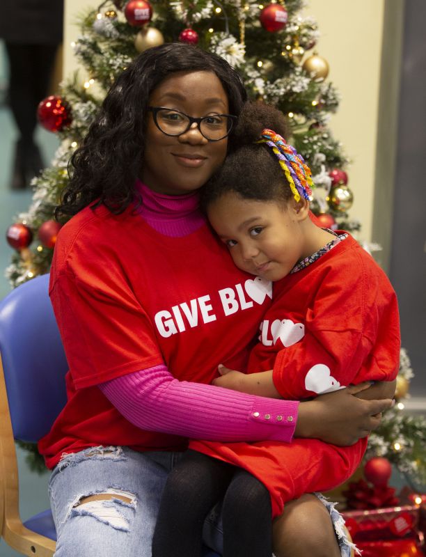 Mum Beth cuddles daughter Evelyn beside a Christmas tree. Both are wearing Give Blood t-shirts. 