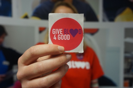 Young lady holding up a 'Give Blood 4 Good' sticker in front of her face